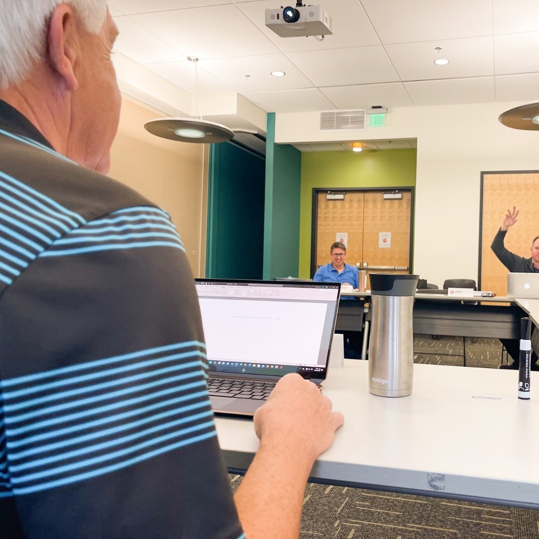 Photo taken from behind of a man sitting in front of a laptop at a conference table while the man across from him raises his hand